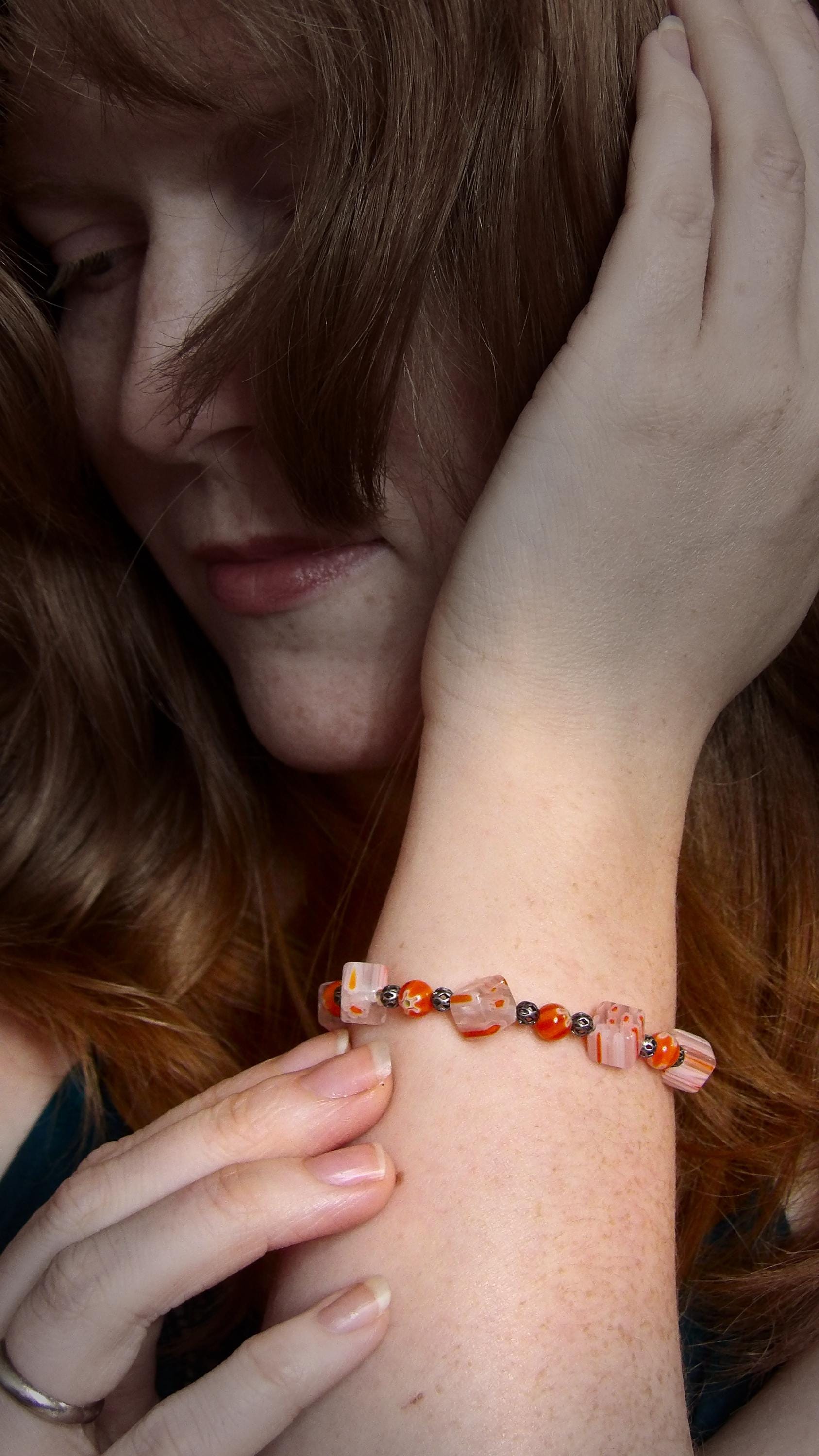 model wearing millefiori glass beaded bracelet made with floral mosaic beads and lace style rounds in a bohemian style. Colors are orange, white and grey.