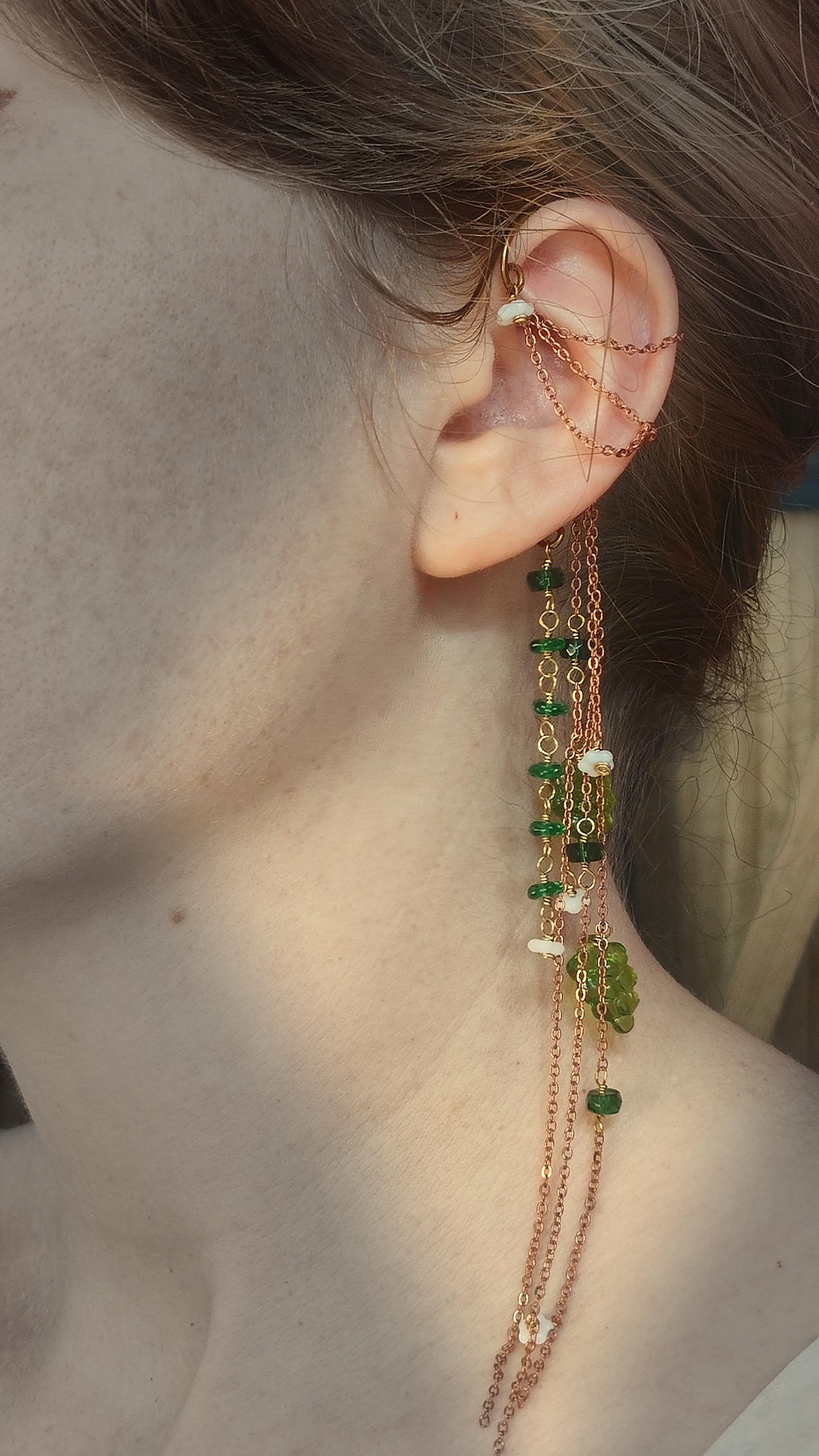 Cottagecore wrap around earring with czech glass flowers and grape beads in shades of green and white. Pierceless design rests behind the ear on a hand forged raw brass frame. Bright copper chains cascade across the top of the ear and from behind the frame for an old world, romantic and rustic vibe. Czech glass beads are hand wrapped and adorned on the the chains throughout the piece