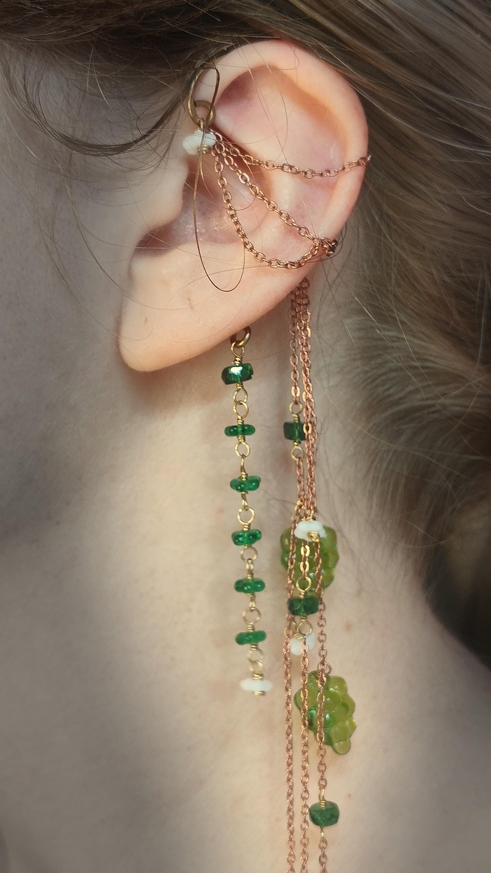 Cottagecore wrap around earring with czech glass flowers and grape beads in shades of green and white. Pierceless design rests behind the ear on a hand forged raw brass frame. Bright copper chains cascade across the top of the ear and from behind the frame for an old world, romantic and rustic vibe. Czech glass beads are hand wrapped and adorned on the the chains throughout the piece