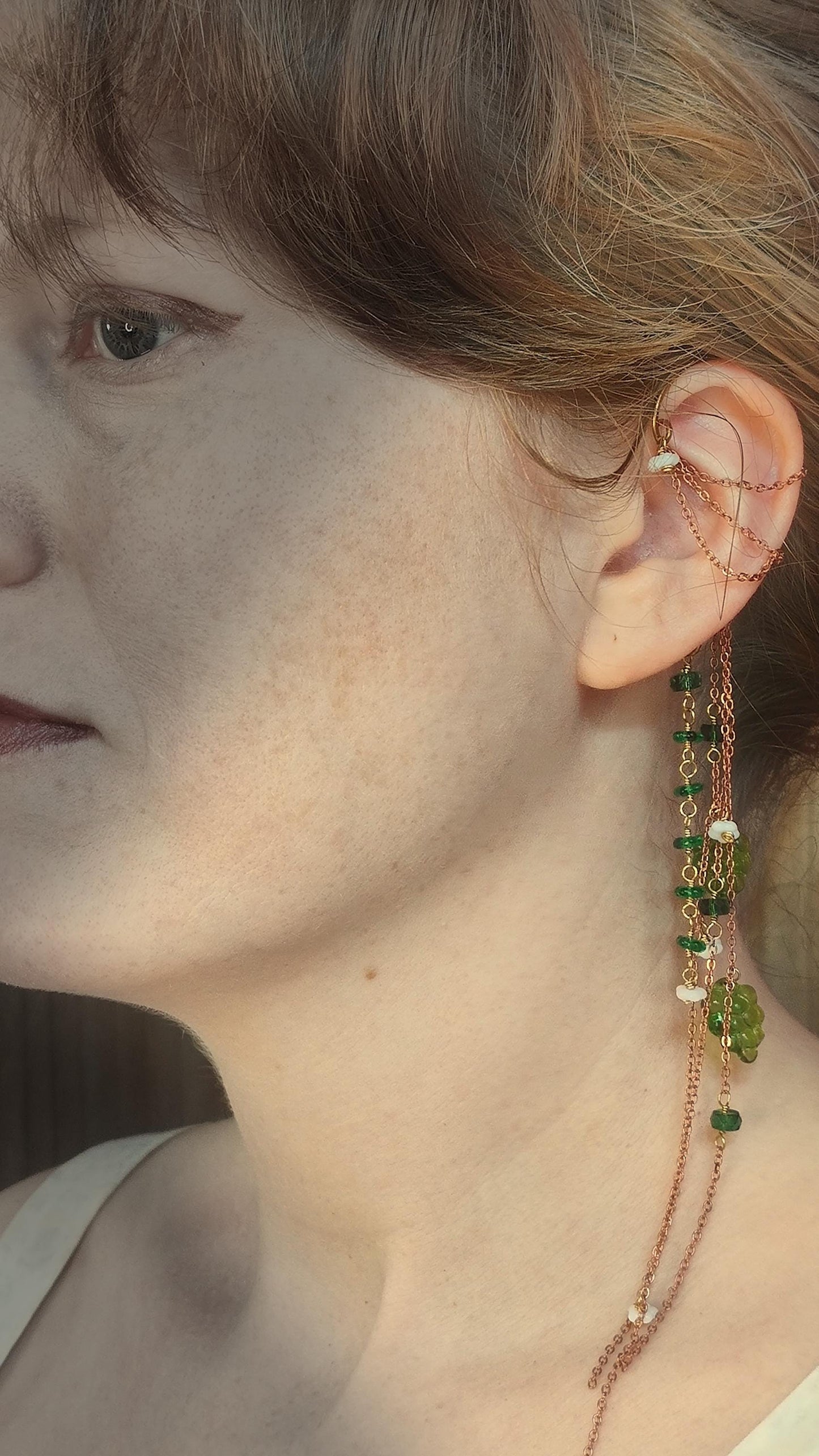Cottagecore wrap around earring with czech glass flowers and grape beads in shades of green and white. Pierceless design rests behind the ear on a hand forged raw brass frame. Bright copper chains cascade across the top of the ear and from behind the frame for an old world, romantic and rustic vibe. Czech glass beads are hand wrapped and adorned on the the chains throughout the piece