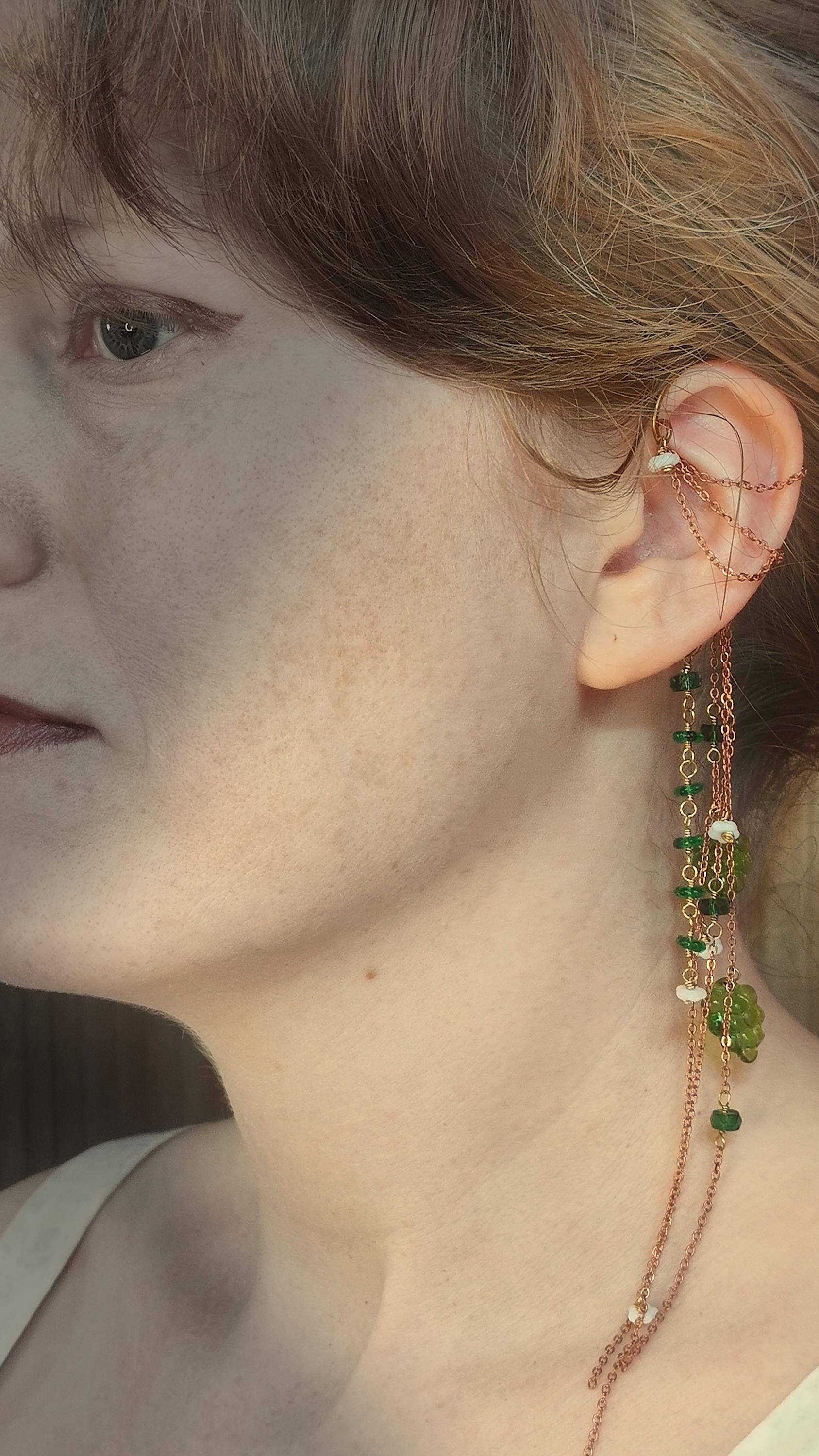 Cottagecore wrap around earring with czech glass flowers and grape beads in shades of green and white. Pierceless design rests behind the ear on a hand forged raw brass frame. Bright copper chains cascade across the top of the ear and from behind the frame for an old world, romantic and rustic vibe. Czech glass beads are hand wrapped and adorned on the the chains throughout the piece