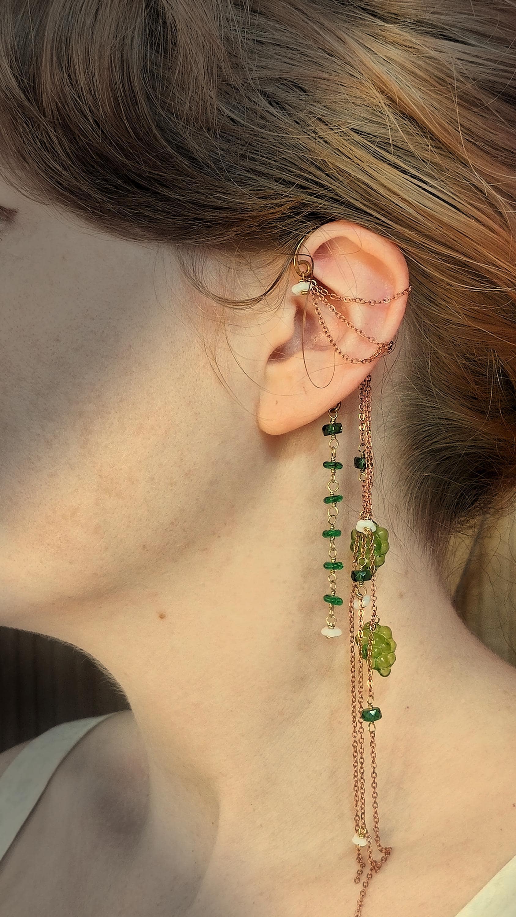 Cottagecore wrap around earring with czech glass flowers and grape beads in shades of green and white. Pierceless design rests behind the ear on a hand forged raw brass frame. Bright copper chains cascade across the top of the ear and from behind the frame for an old world, romantic and rustic vibe. Czech glass beads are hand wrapped and adorned on the the chains throughout the piece
